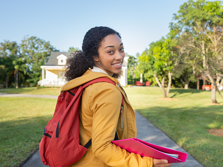 young woman walking down a sidewalk, head turned to face the camera, holding a notebook binder and backpack, green grass and trees and a house structure in the background