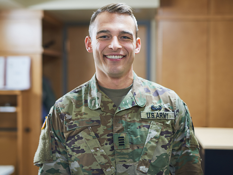 smiling young man wearing US Army camouflage uniform, facing the camera, indistinct wooden cabinetry in the background